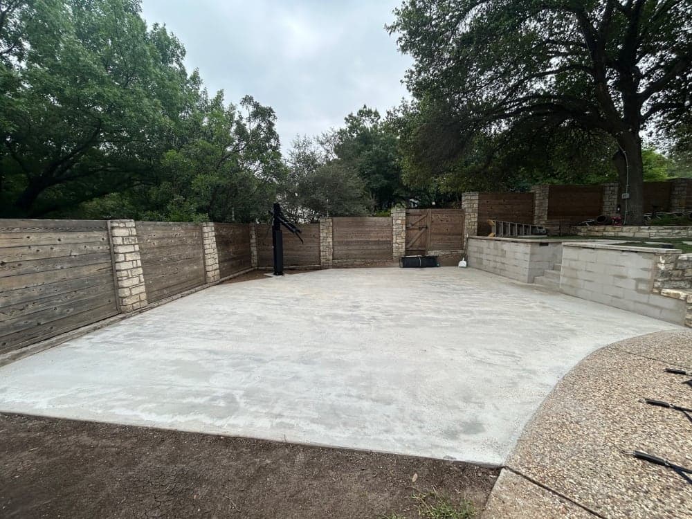 Bare concrete pad in a stone-and-wood-fenced backyard before basketball court surfacing in Austin, Texas