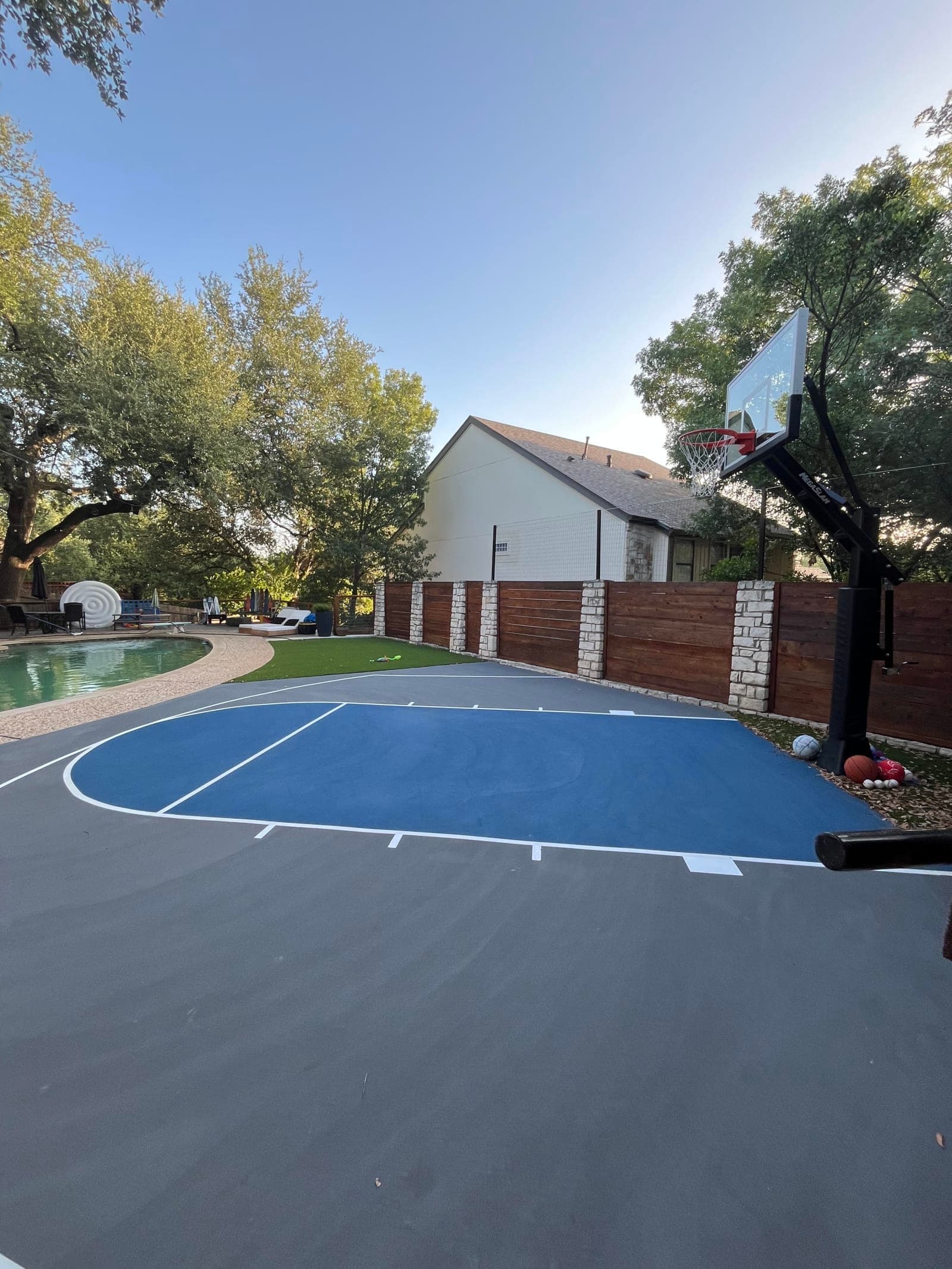 Finished blue and gray backyard basketball court next to a pool with a stone and wood fence in Austin, Texas